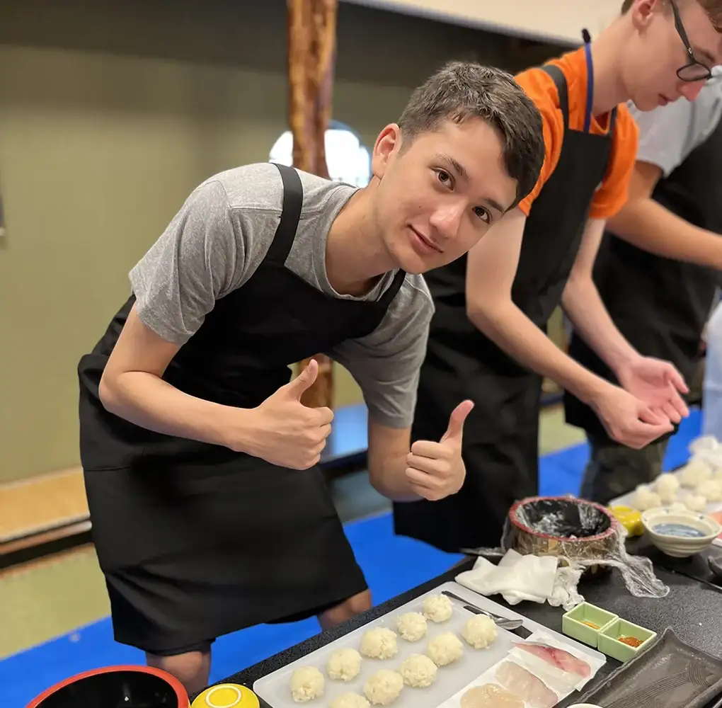 Young man in an apron smiling and giving thumbs up while preparing sushi rice balls in a Japanese cooking class